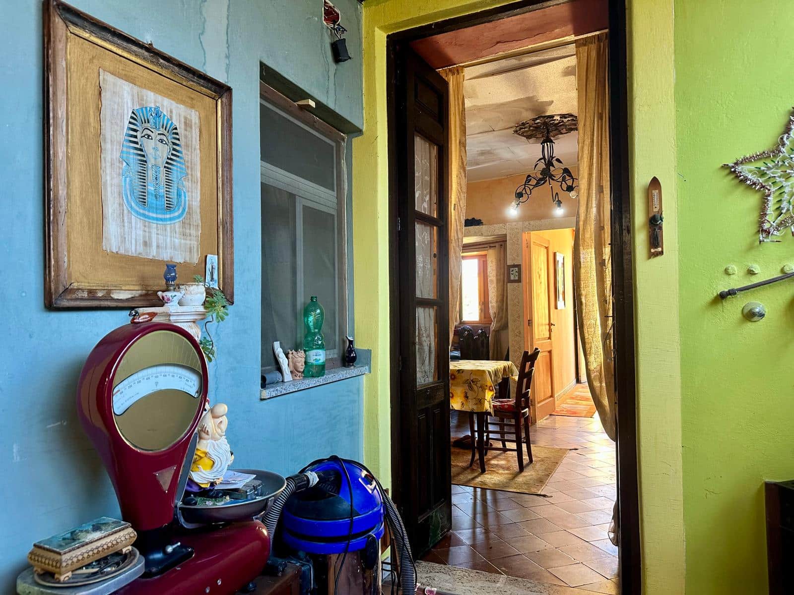 Foto di un interno di casa con pareti colorate, stile eclettico, e decorazioni vintage. Viene visualizzata una zona cucina e soggiorno, con dettagli come quadri, oggetti d'antiquariato e un aspirapolvere tradizionale, creando un ambiente caloroso e accogliente.