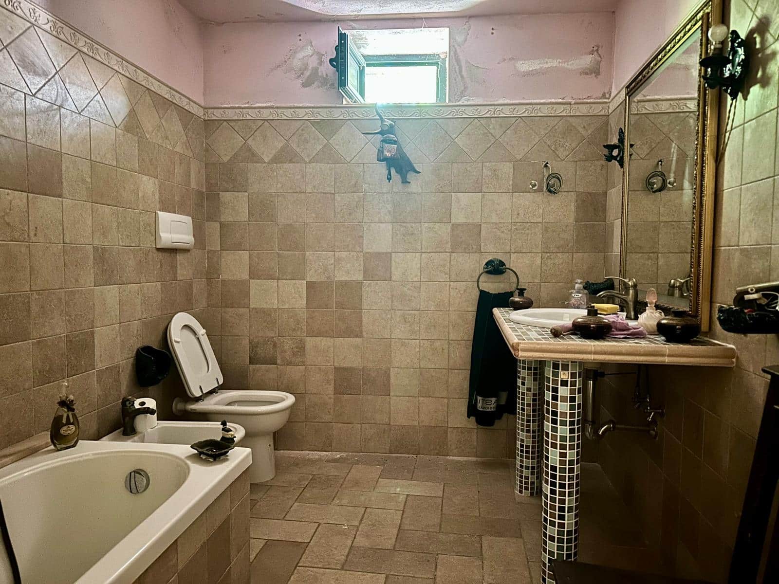 Bagno rustico con pareti e pavimenti in piastrelle beige, vasca, WC, lavabo con specchio grande, finestra piccola aperta, decorazioni vintage e dettagli in mosaico con stile classico e accogliente.