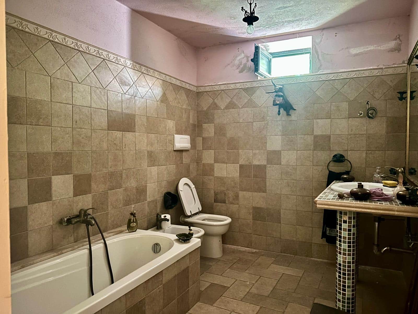 Bagno rustico con vasca, WC e lavabo con piastrelle beige e parete decorativa, finestra piccola per ventilazione naturale, stile vintage e arredi in ceramica e metallo.