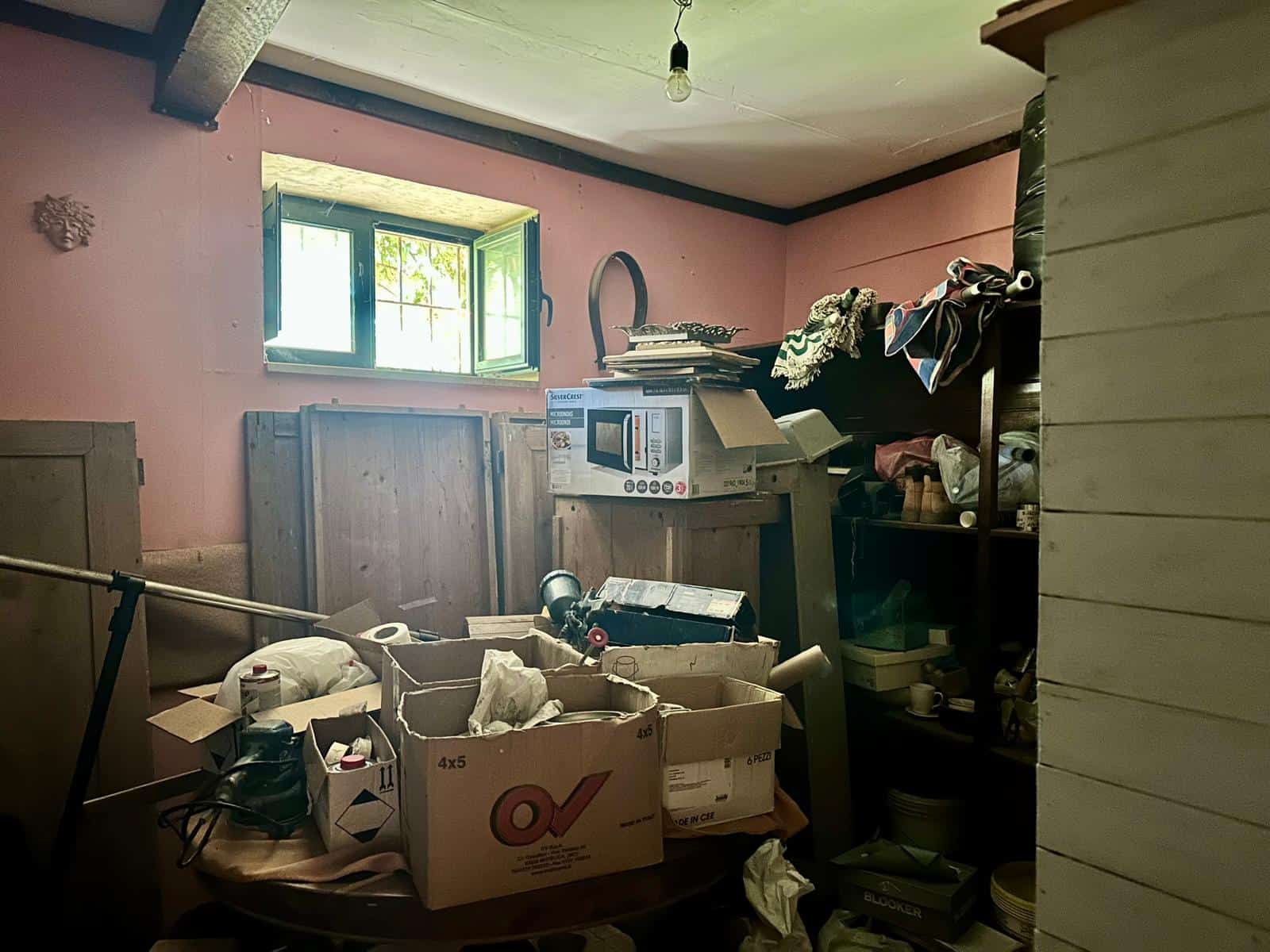 Spazio disordinato con scatole e oggetti vari in una stanza con pareti rosa e finestra aperta, suggerendo un'area di deposito o svalutazione di interni.