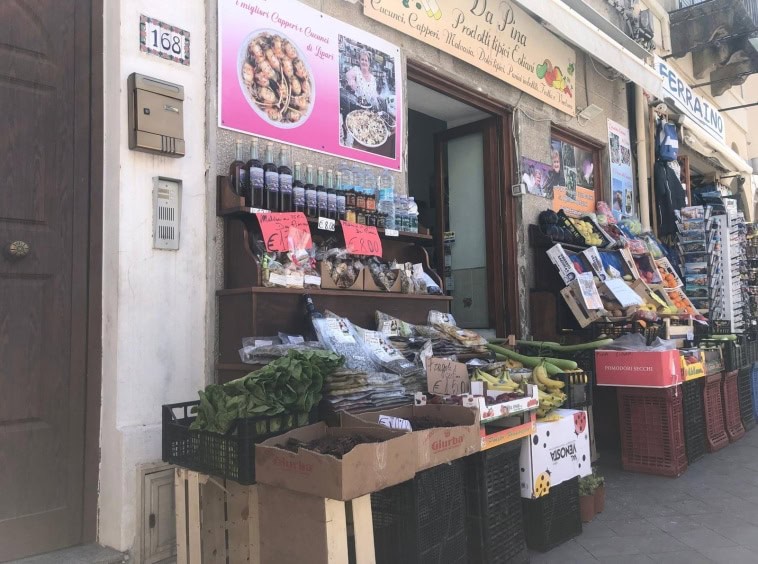 Prezzo di spesa di prodotti freschi e locali, frutta e verdura all'aperto, negozio di alimentari tradizionale, con scatole di pomodori secchi, banane, foglie di insalata, e bottiglie di vino o olio, in Italia, vicino alla casa di NextCasa.