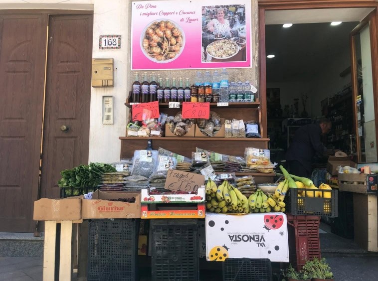 Pomodoro di frutta e verdura fresche in vendita davanti a un negozio di alimentari in Italia. Banco di prodotti con banane, insalate, spezie e altri alimenti locali, in un quartiere urbano.