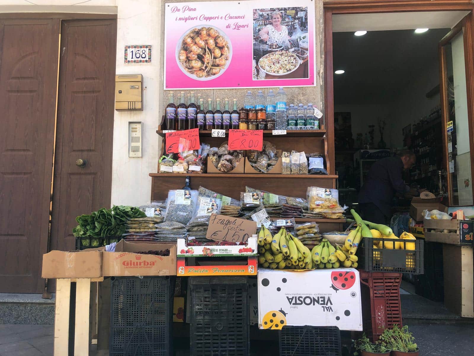 Pomodoro di frutta e verdura fresche in vendita davanti a un negozio di alimentari in Italia. Banco di prodotti con banane, insalate, spezie e altri alimenti locali, in un quartiere urbano.