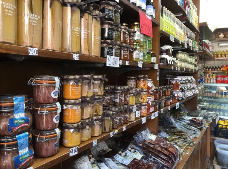 Barattoli di conserve e prodotti alimentari rappresentati in un negozio di alimentari, con scaffali pieni di merce di vario tipo e prezzi visibili.