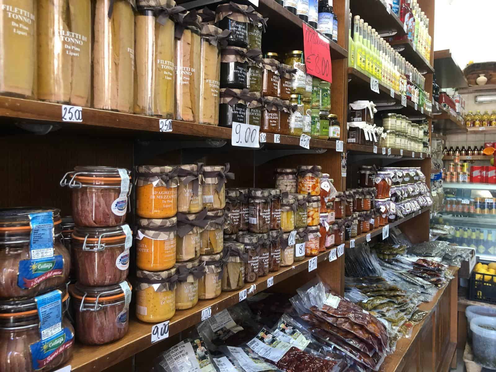 Barattoli di conserve e prodotti alimentari rappresentati in un negozio di alimentari, con scaffali pieni di merce di vario tipo e prezzi visibili.