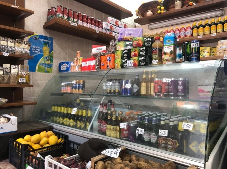 Pompe di frutta e prodotti alimentari dietro il banco di una bottega alimentare, con scaffali di conserve e bevande, evidenziando l'offerta di cibi freschi e confezionati, ideale per negozi di alimentari e servizi di consegna pasti.