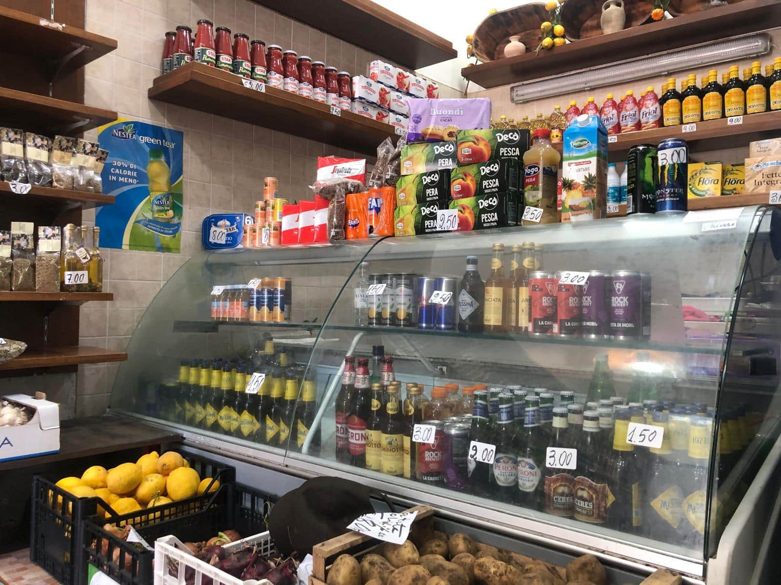 Pompe di frutta e prodotti alimentari dietro il banco di una bottega alimentare, con scaffali di conserve e bevande, evidenziando l'offerta di cibi freschi e confezionati, ideale per negozi di alimentari e servizi di consegna pasti.