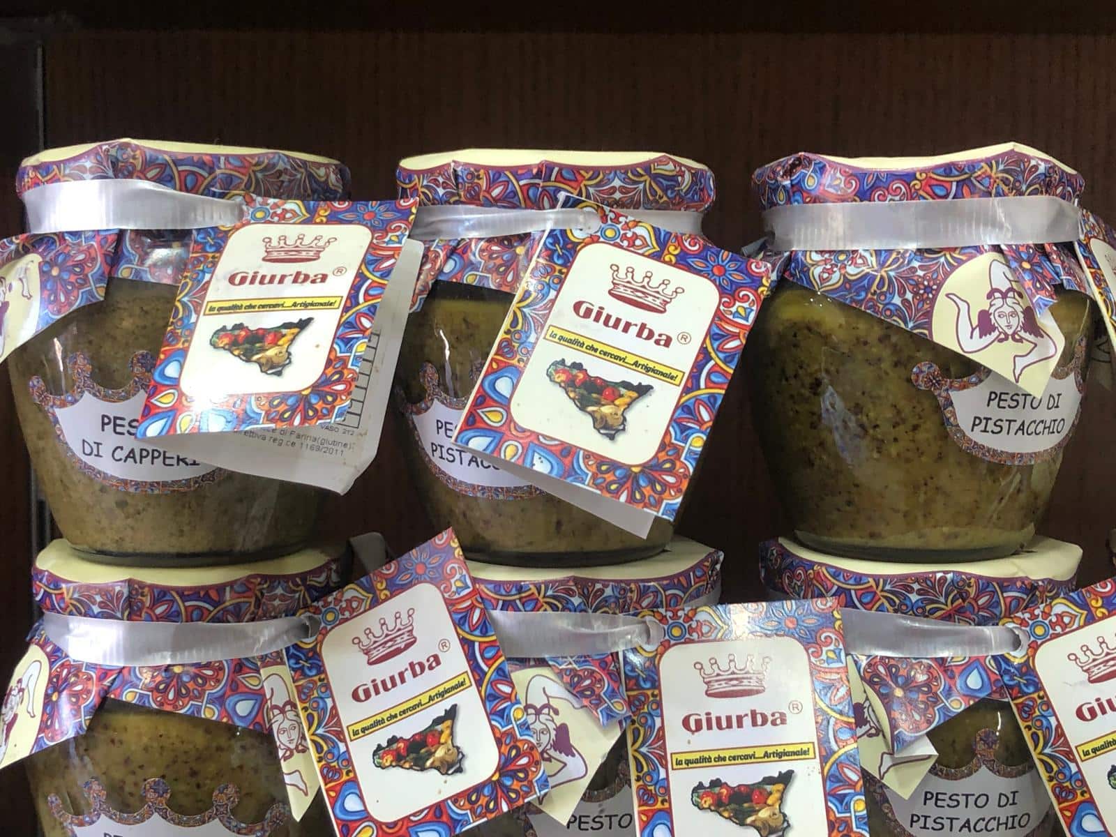 Pesto di pistacchio Giurba, prodotto artigianale di alta qualità, confezionato in vasetti decorati con etichette colorate e disegni tradizionali siciliani, ideale per ricette gourmet e antipasti raffinati.