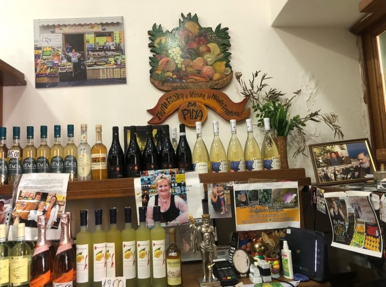 Bottiglie di vino e liquori in esposizione nel negozio Da Pina, con decorazioni, foto e volantini pubblicitari, che evidenziano i prodotti tipici colorati e autentici, rappresentando un punto vendita di specialità alimentari locali.