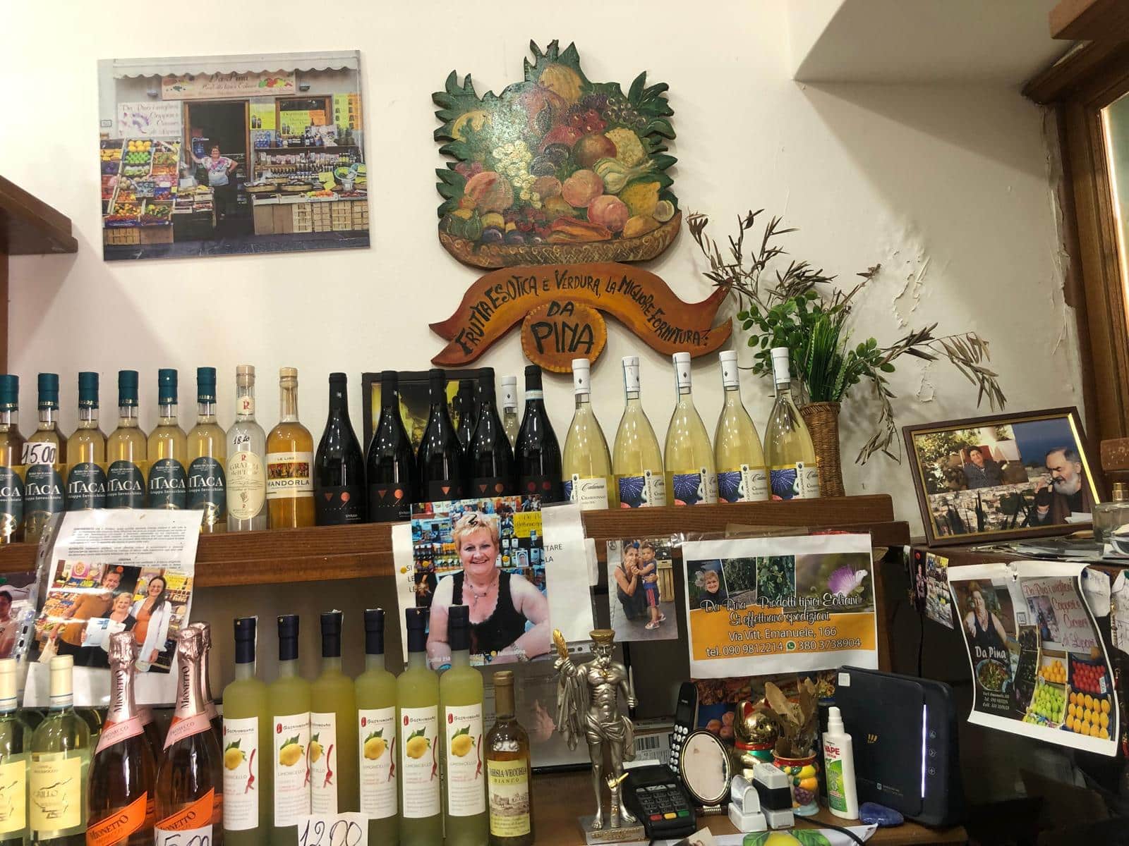 Bottiglie di vino e liquori in esposizione nel negozio Da Pina, con decorazioni, foto e volantini pubblicitari, che evidenziano i prodotti tipici colorati e autentici, rappresentando un punto vendita di specialità alimentari locali.