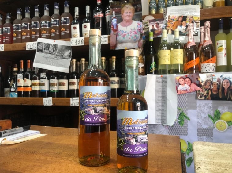 Bottiglie di liquore alla fira da Pina con etichette colorate, in primo piano, all’interno di una bottega di prodotti regionali siciliani; sfondo con scaffali di vino e confet .