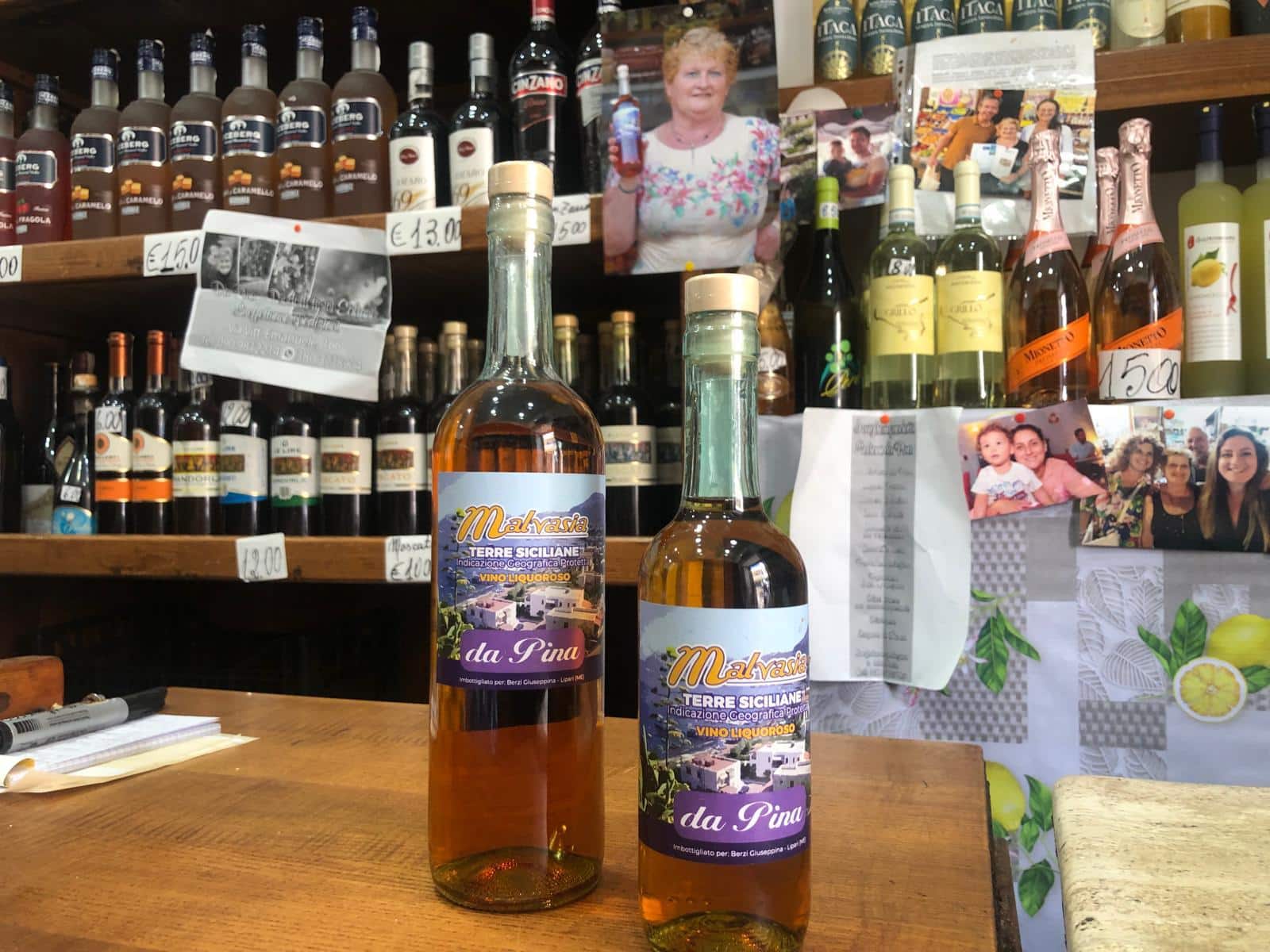 Bottiglie di liquore alla fira da Pina con etichette colorate, in primo piano, all’interno di una bottega di prodotti regionali siciliani; sfondo con scaffali di vino e confet .
