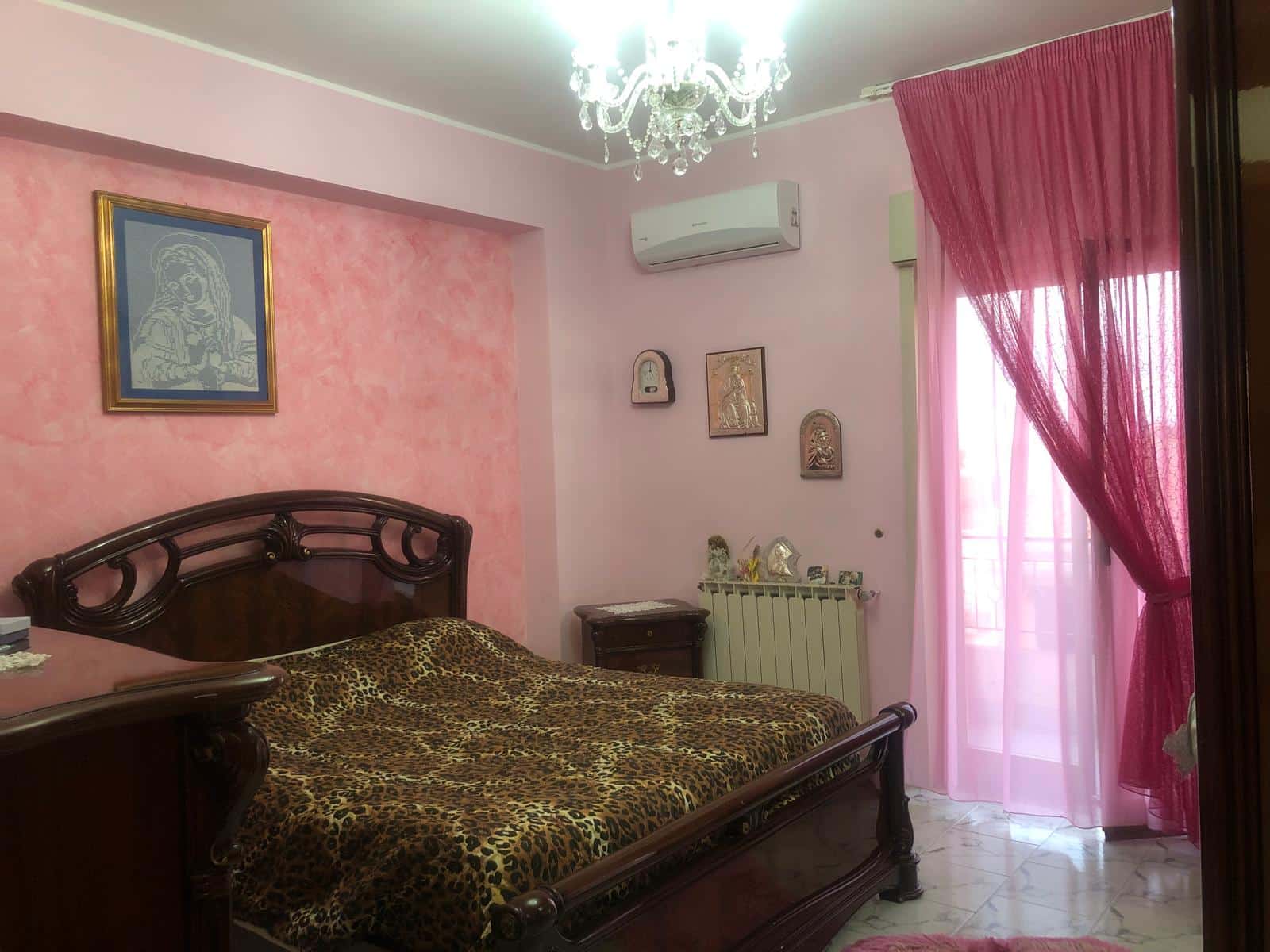 Letto matrimoniale in stile classico con lenzuola a stampa animale, pareti rosa, decorazioni religiose e tendaggi rosa trasparenti su finestra e porta balcone, aria condizionata e lampadario chandelier.