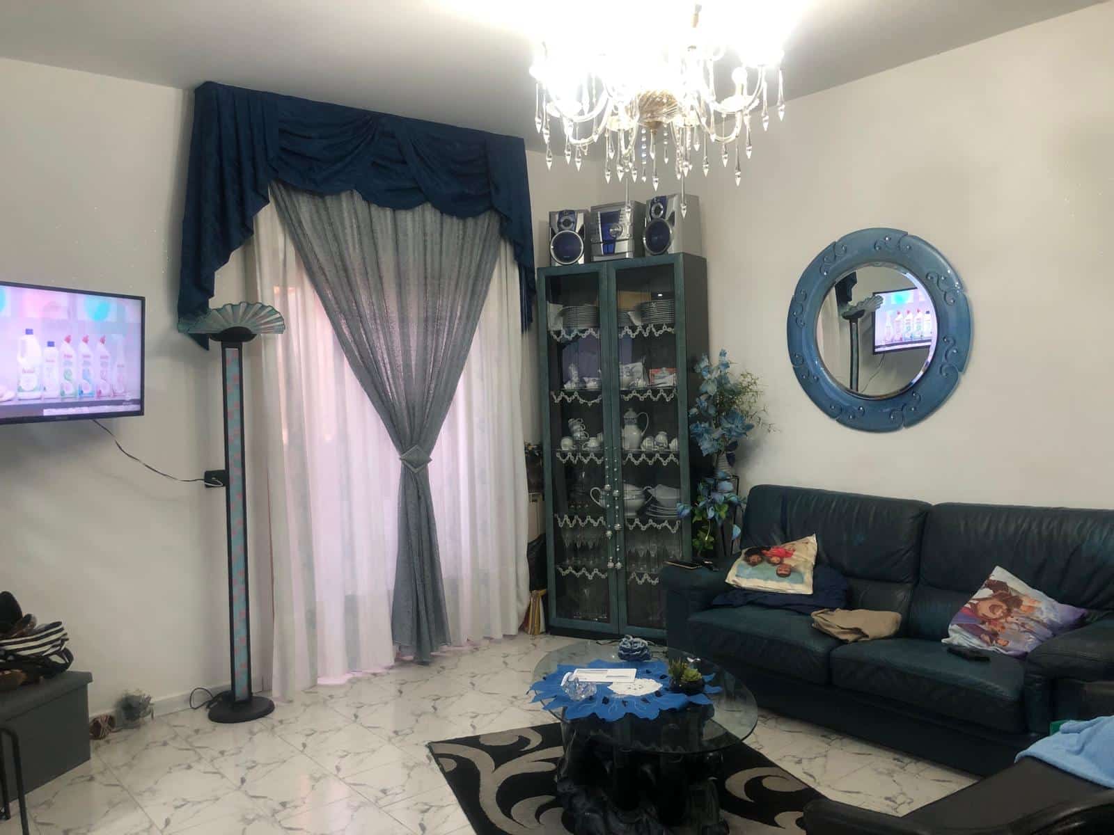 Composizione di soggiorno con divano in pelle nera, tavolino in vetro, specchio decorativo blu sulla parete, mobile vetrina con piatti, satellite e TV, tende con drappeggio blu e beige, lampada a piantana e lampadario elegante.