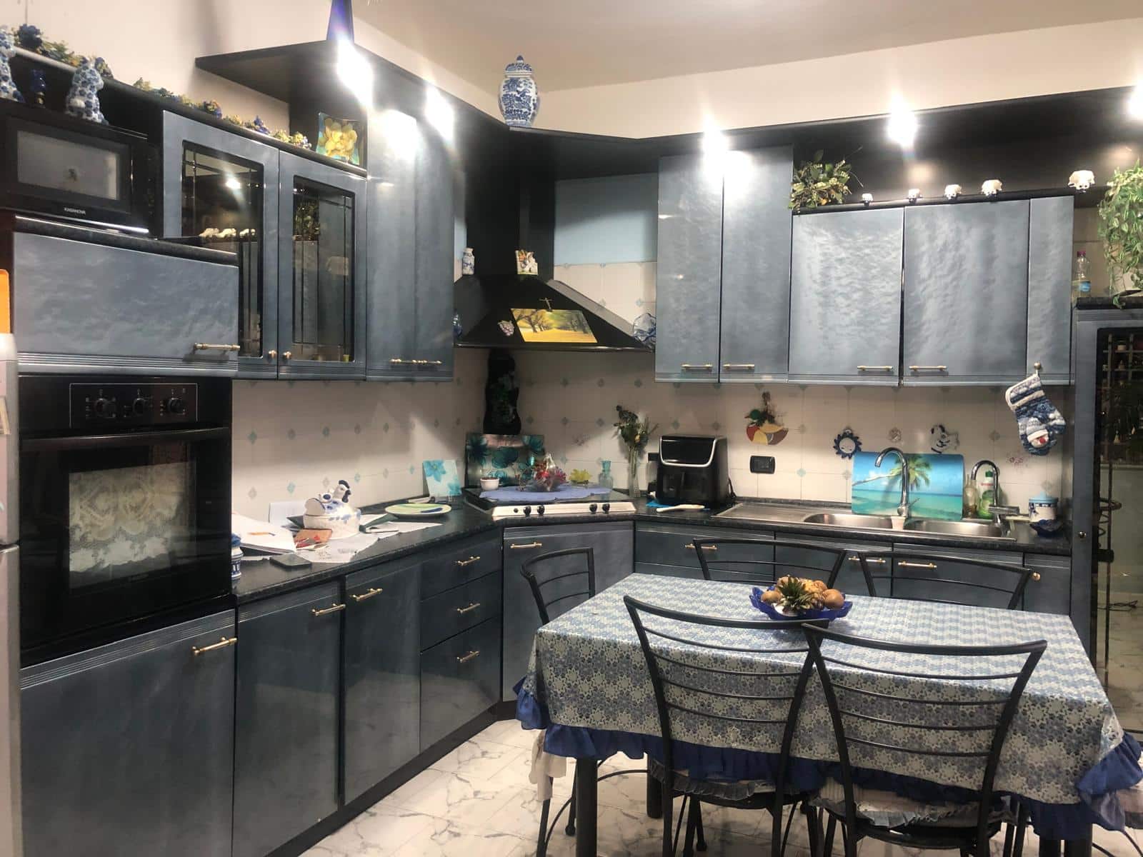 Cucina moderna con mobili blu scuro e tavolo da pranzo con tovaglia floreale, elettrodomestici neri e decorazioni colorate, ambiente accogliente e ben illuminato, stile classico e funzionale.