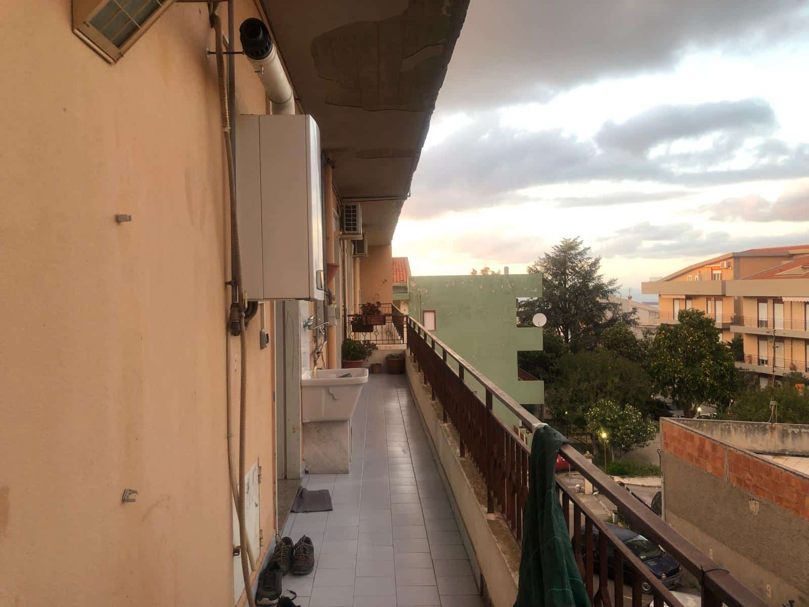 Balcone ampio e luminoso in un appartamento con vista sui tetti e sul cielo nuvoloso, con piante, pavimento in piastrelle e alcuni elettrodomestici, ideale per relax e vivere all'aperto.