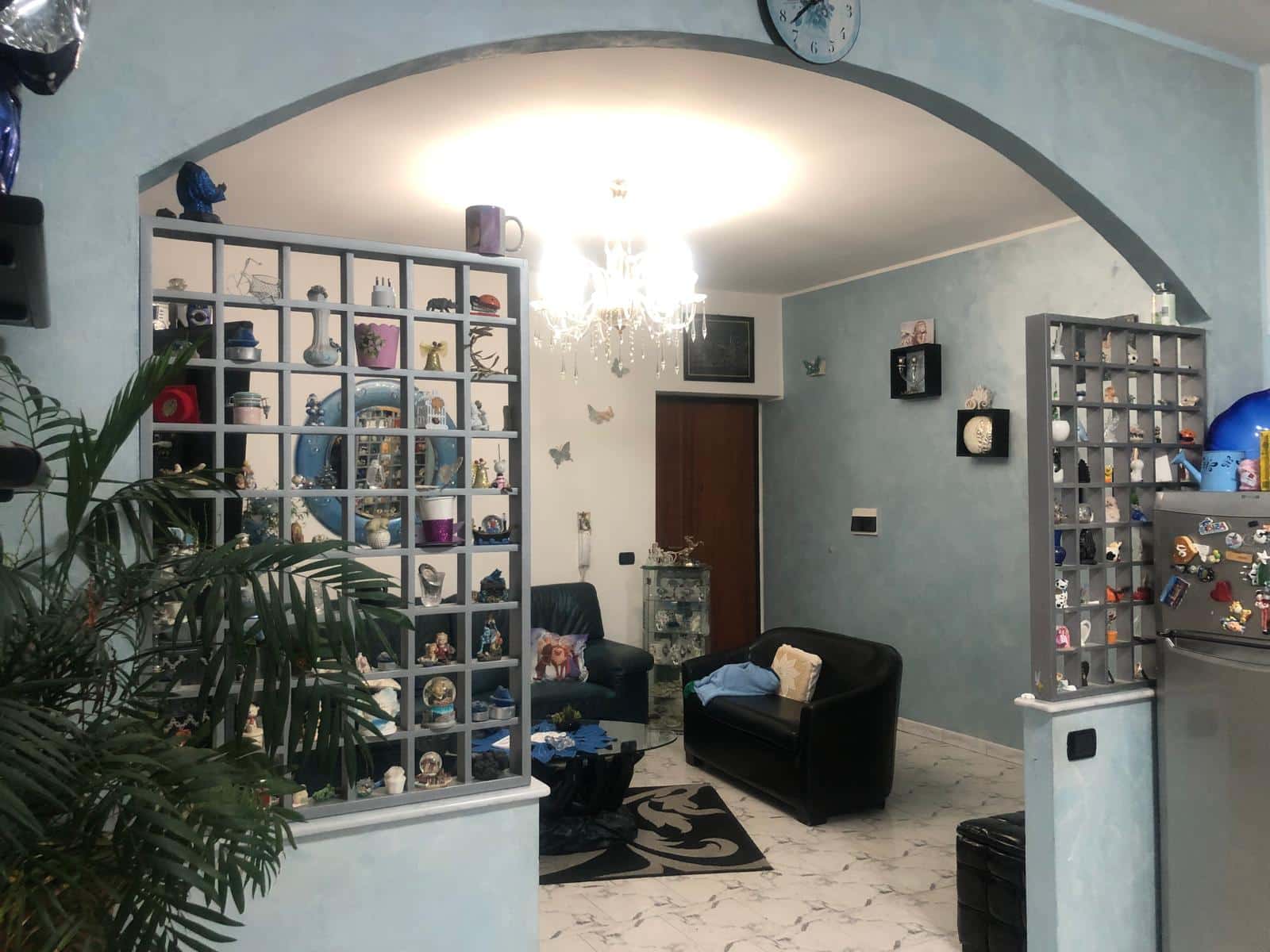 Vetrina decorativa con oggetti d'arredo e accessori per la casa in un soggiorno con divani neri e pareti azzurro chiaro, illuminato da lampadario elegante, perfetta per interni moderni.