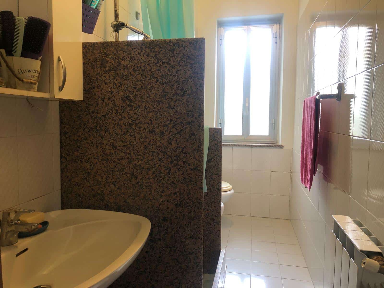 Lavandino e termosifone in bagno con finestra, pareti piastrellate e accessori da bagno in esposizione, ambiente luminoso e funzionale, ideale per servizi di prossimità e comfort domestico.