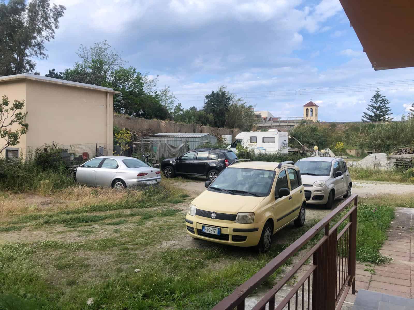 Auto a noleggio in cortile con altri veicoli, con sfondo di case, natura e colline, vista di un paesaggio rurale.