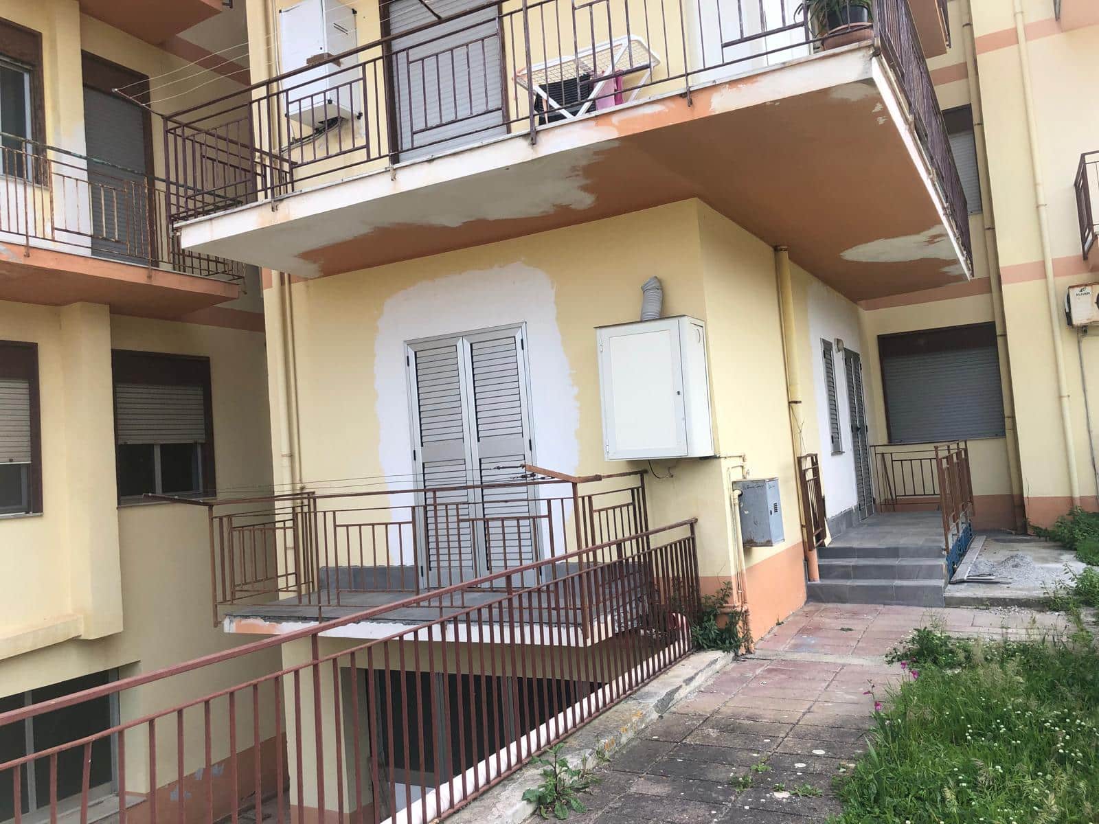 Balcone di appartamento in fase di ristrutturazione con ringhiera in metallo, scala di accesso esterna in pietra, pareti dipinte e spazio verde annesso, in contesto condominiale in città.