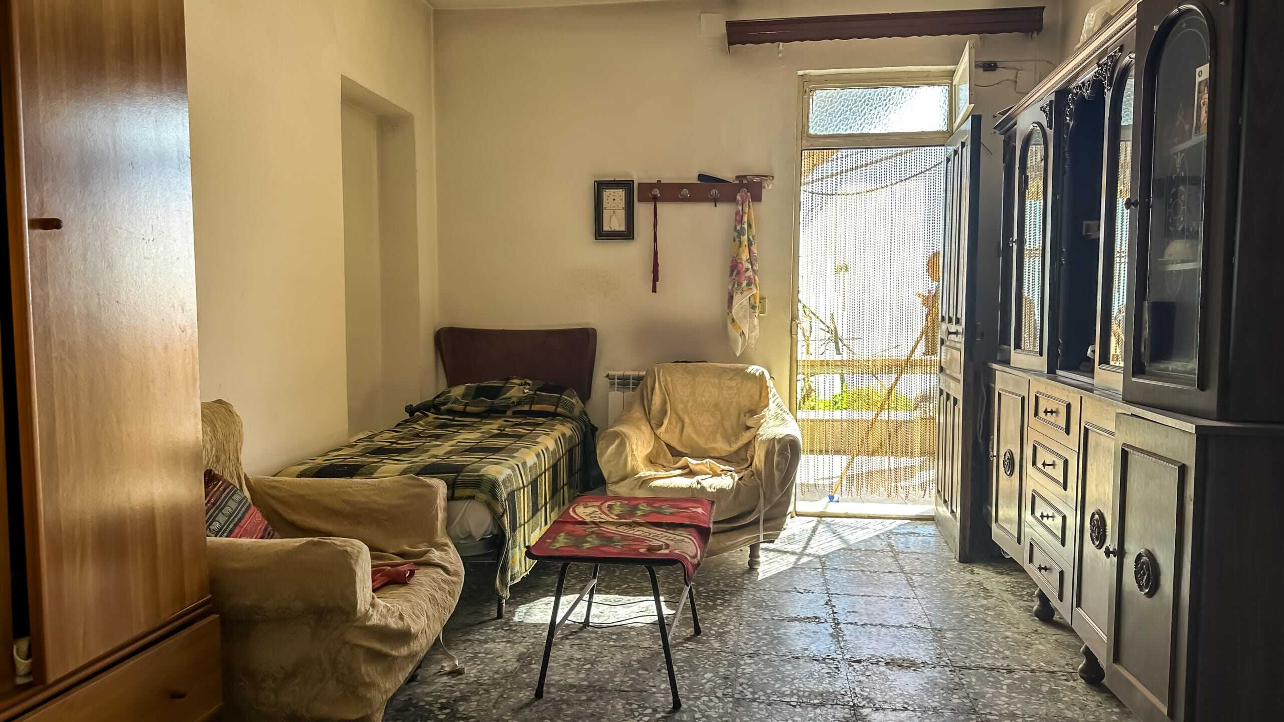 Stanza soggiorno con divano, poltrona, armadio in legno e porta finestra con luce naturale, ambiente semplice e accogliente.