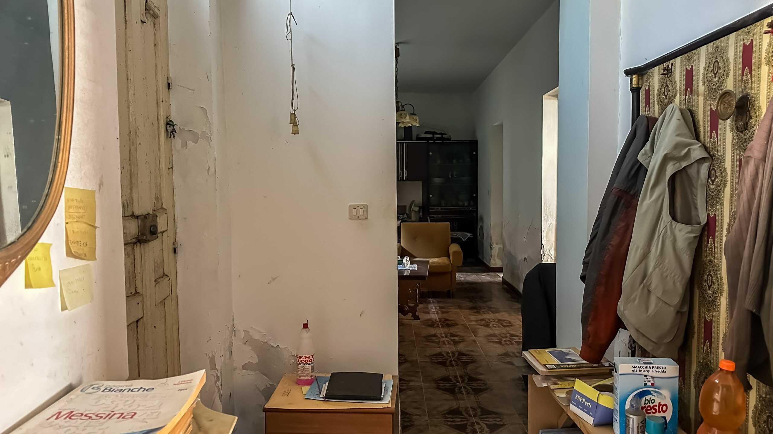 Soggiorno con arredi vintage, pareti da ristrutturare e pavimenti in maiolica, ambiente abitativo con oggetti e vestiti appesi, atmosfera di casa da rinnovare, immagini di interni di abitazioni da sistemare, NextCasa immobiliar.