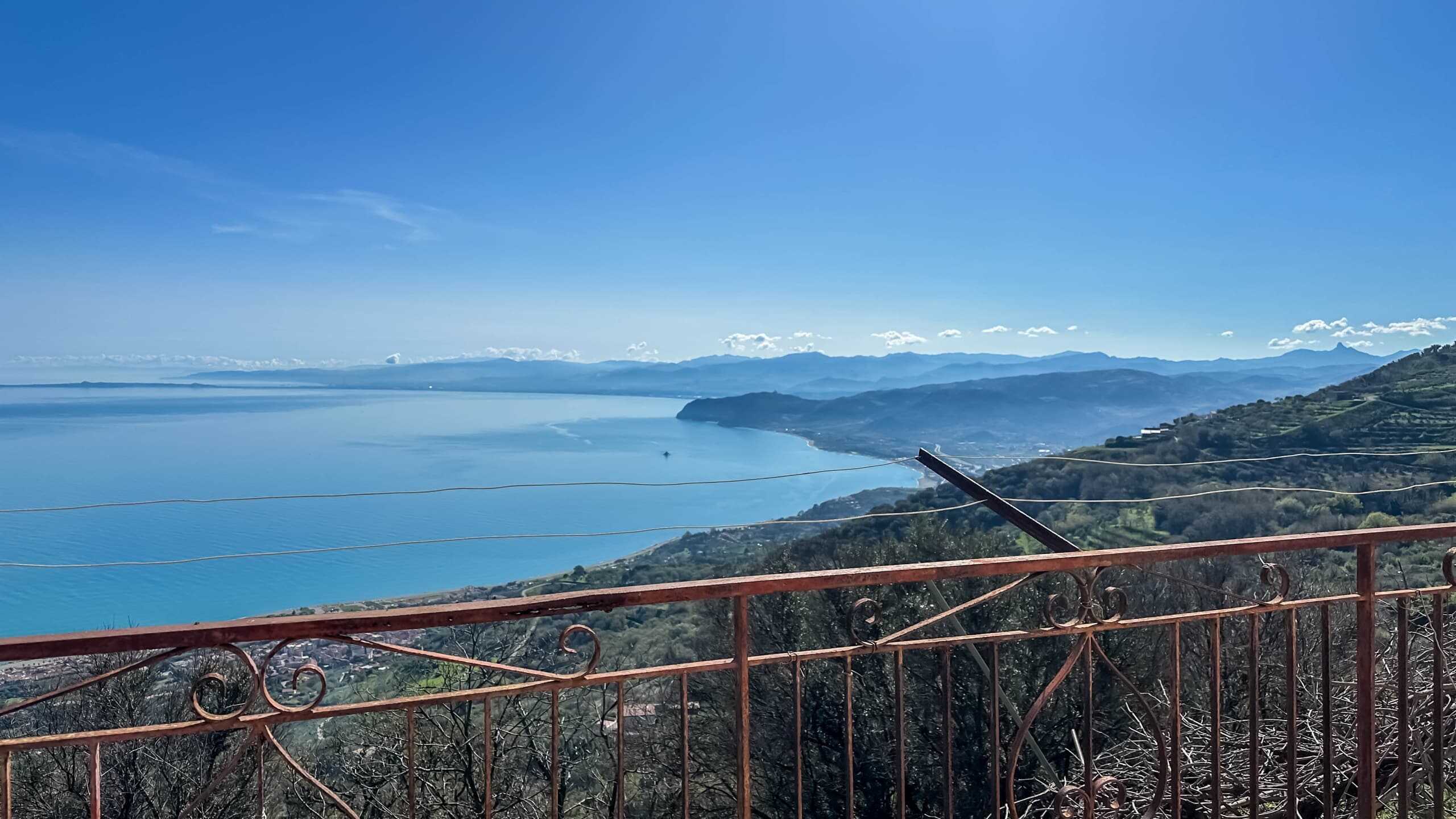 VISTA PANORAMICA DELLA COSTA E DEL MARE DAL TERRAZZO CON BARRIERA IN FERRO VINTAGE, SPETTACOLARE PANORAMICA SULLO SPAZIO AZZURRO DEL MARE E SULLA NATURA VERDE DELLE COLLINE CIRCOSTANTI, CON SFONDO MONTAGNOSO E CIELO SERENO.
