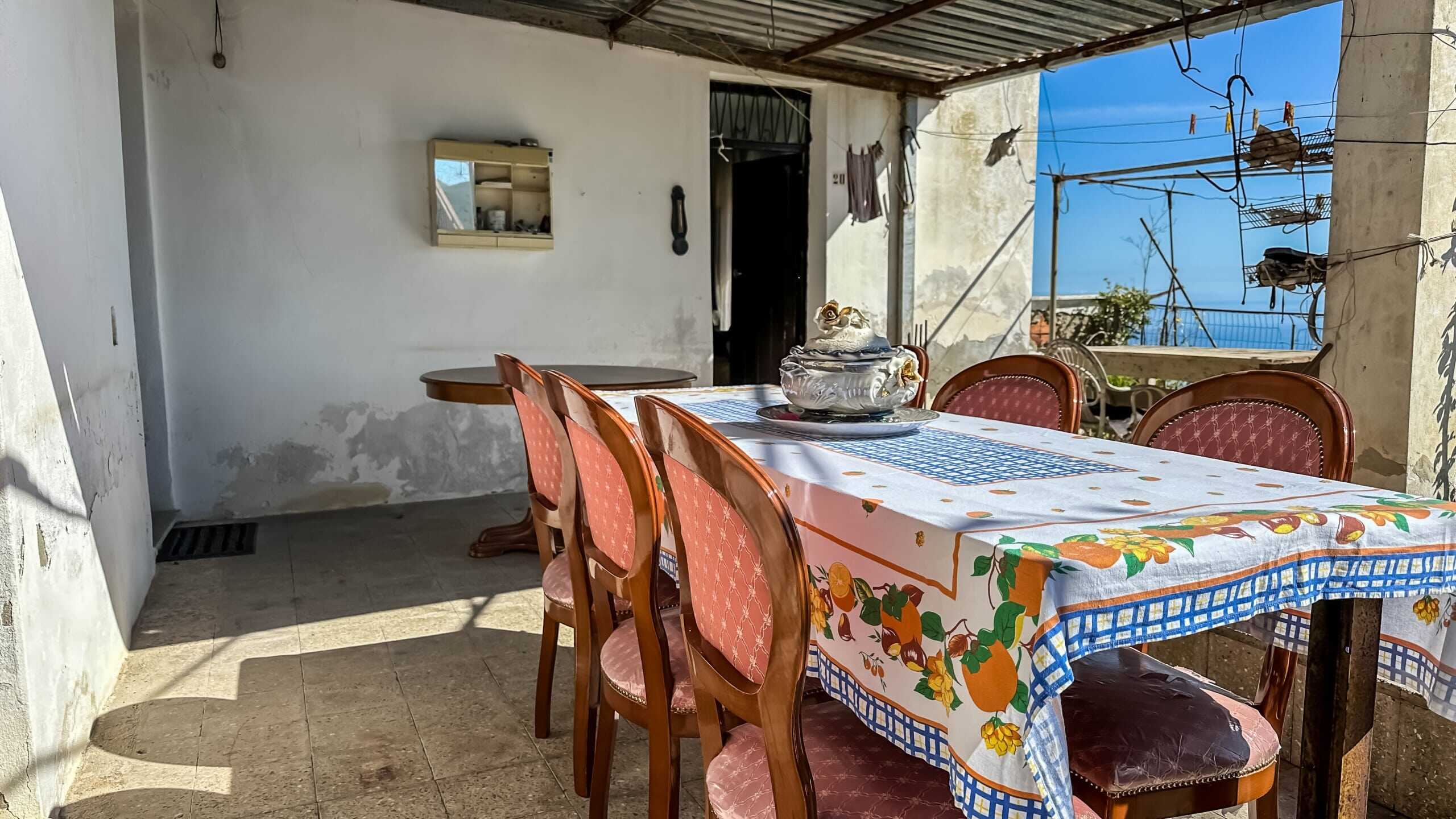 Tavolo da pranzo con tovaglia colorata, sedie in velluto rosa, terrazza con vista sul mare, arredi rustici, spazio esterno, ambiente accogliente, casa al mare, NextCasa, proprietà immobiliare.