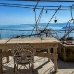VISTA PANORAMICA: terrazza con vista sul mare e sulle montagne, arredamento rustico con tavolo in legno naturale e sedia in vimini, atmosfera tranquilla e suggestiva, spazio esterno con piante e stile autentico.