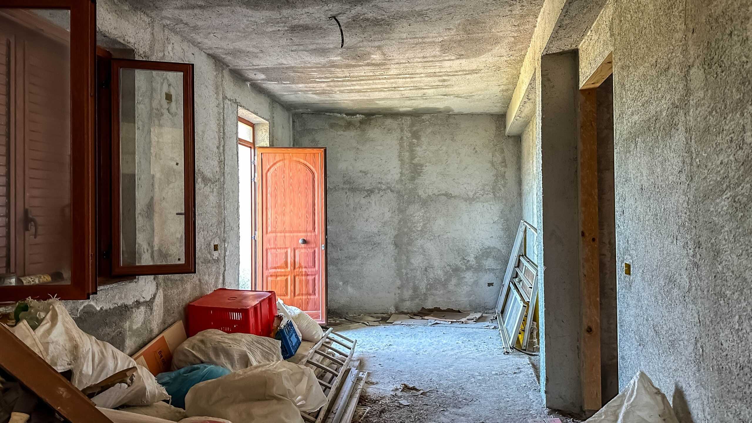 Stanza in costruzione con pareti grezze, infissi in legno e porta d’ingresso in stile classico, pianoterra aperto, spazio di lavoro in fase di rinnovamento per una futura abitazione.