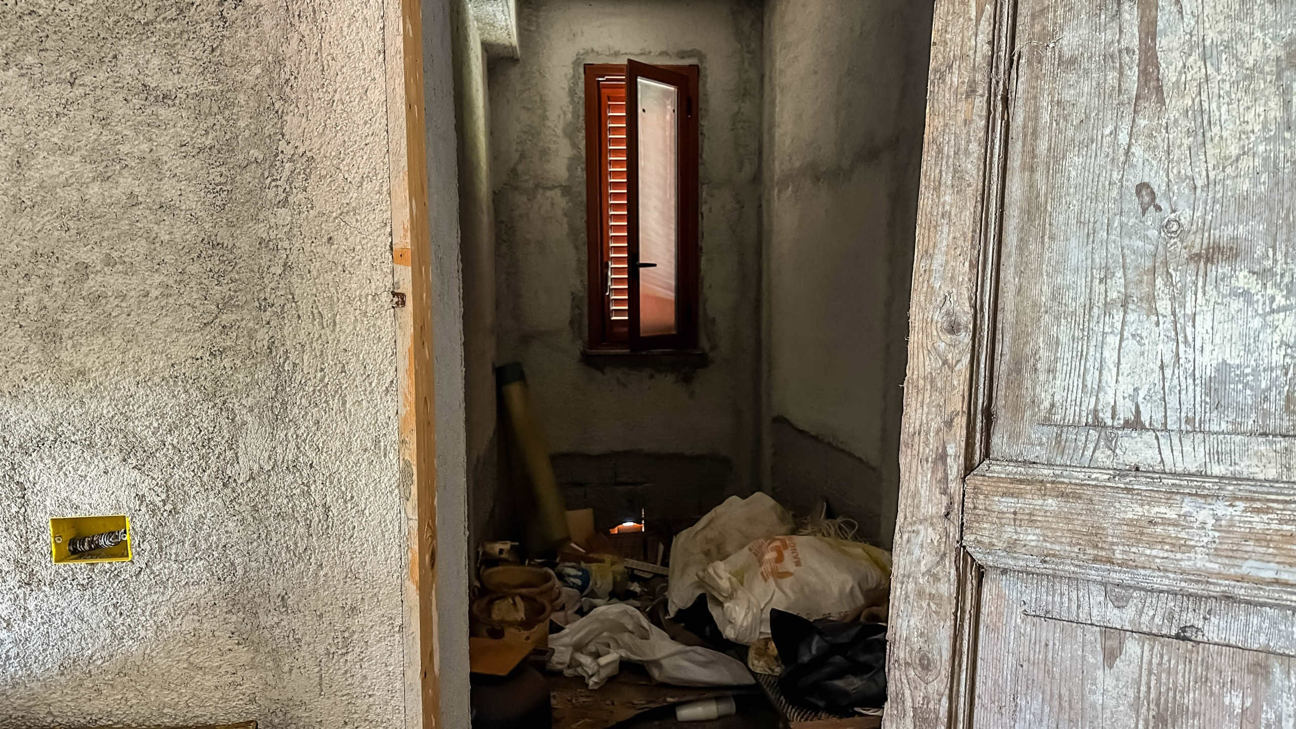 Ripresa di un interno di casa in fase di ristrutturazione, con pareti in intonaco grezzo, finestra con tapparella e pavimento trascurato, mostrando lo stato attuale di un intervento edilizio.