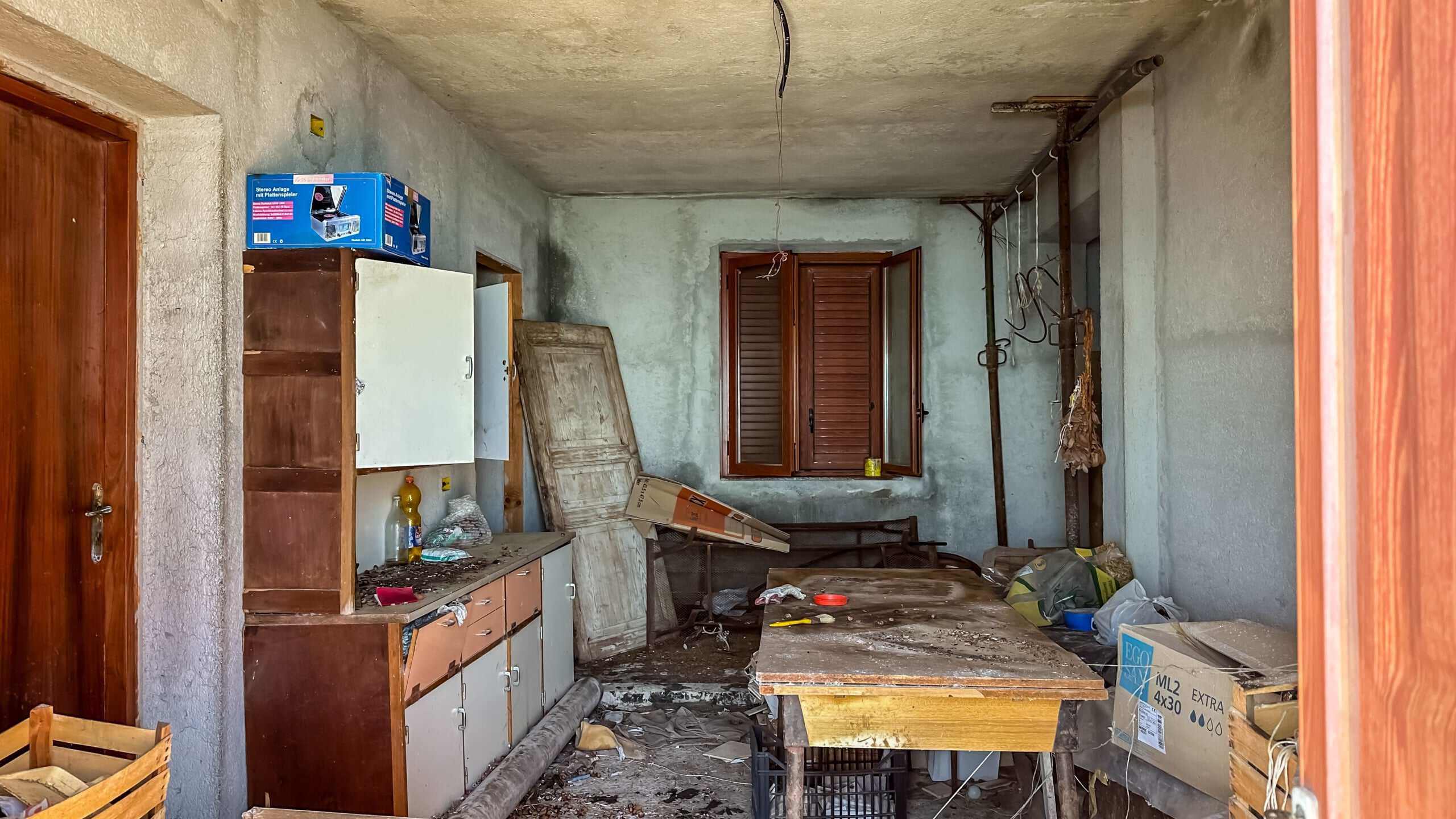 Vecchia cucina disordinata, mobili deteriorati e finestra con tapparelle di legno, in un ambiente di casa abbandonata o in ristrutturazione, con evidenti segni di degrado e sporco.