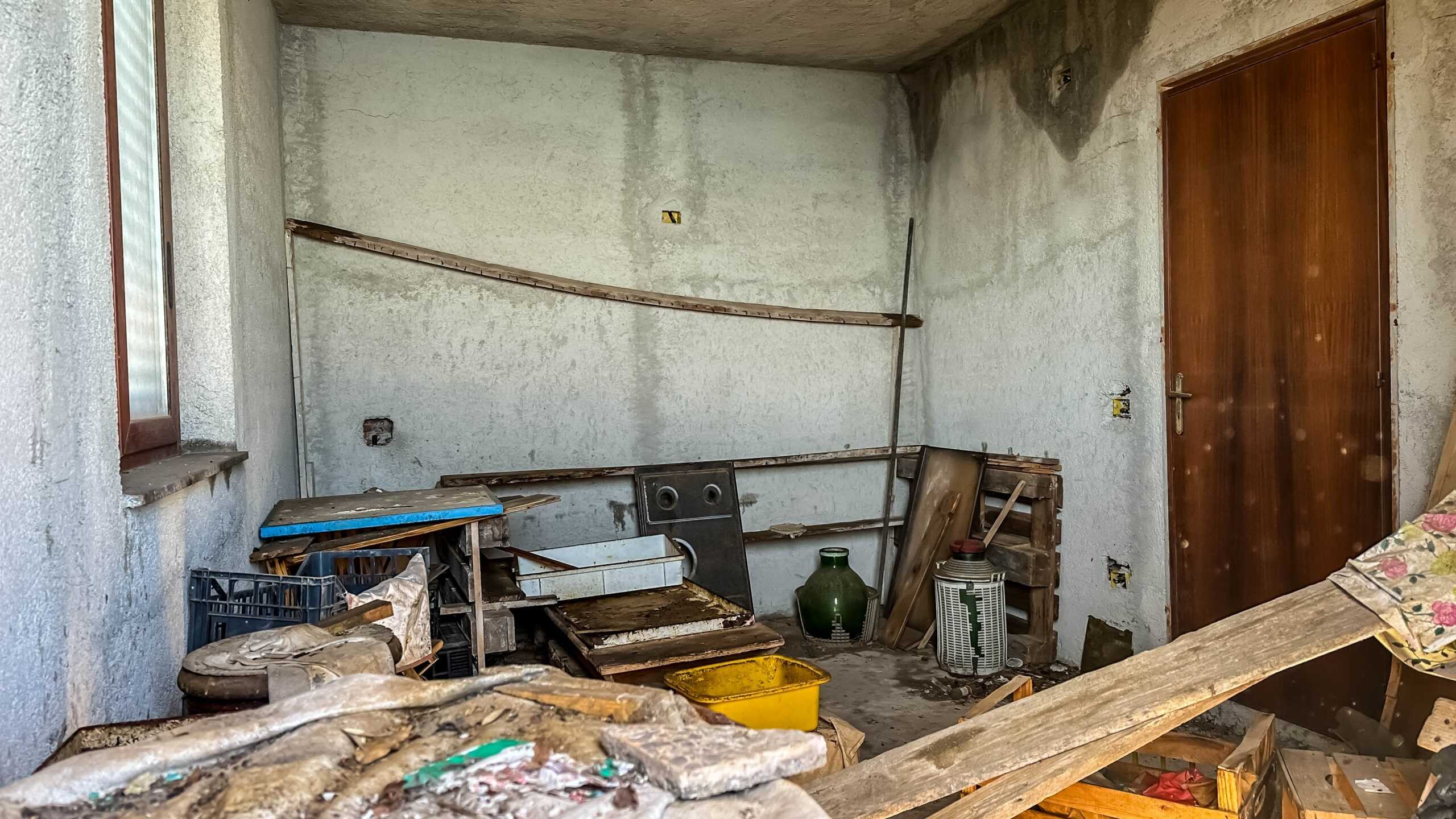 Fondo con pareti di calcestruzzo e porta in legno massello peso, ambiente in stato di ristrutturazione, scatole di legno e oggetti da lavoro, spazio di lavoro trasandato e in fase di rinnovamento di una casa NextCasa.