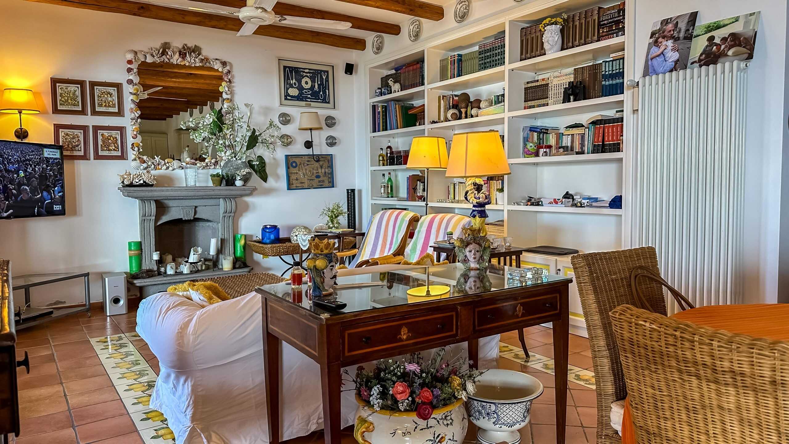 Soggiorno elegante con libreria, camino in pietra, divani con coperte colorate, illuminate da lampade gialle, decorazioni floreali, TV e pareti decorate, atmosfera calda e accogliente, ideale per vivere momenti di relax e convivialità.