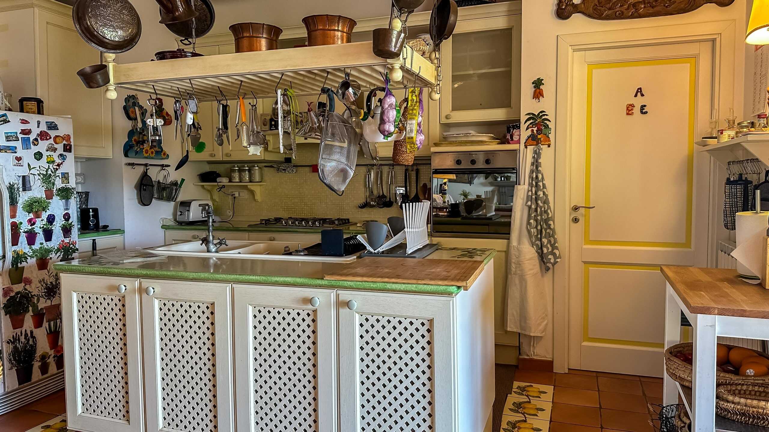 Pentola e utensili da cucina appesi al soffitto in cucina colorata con mobili bianchi e superficie in legno.