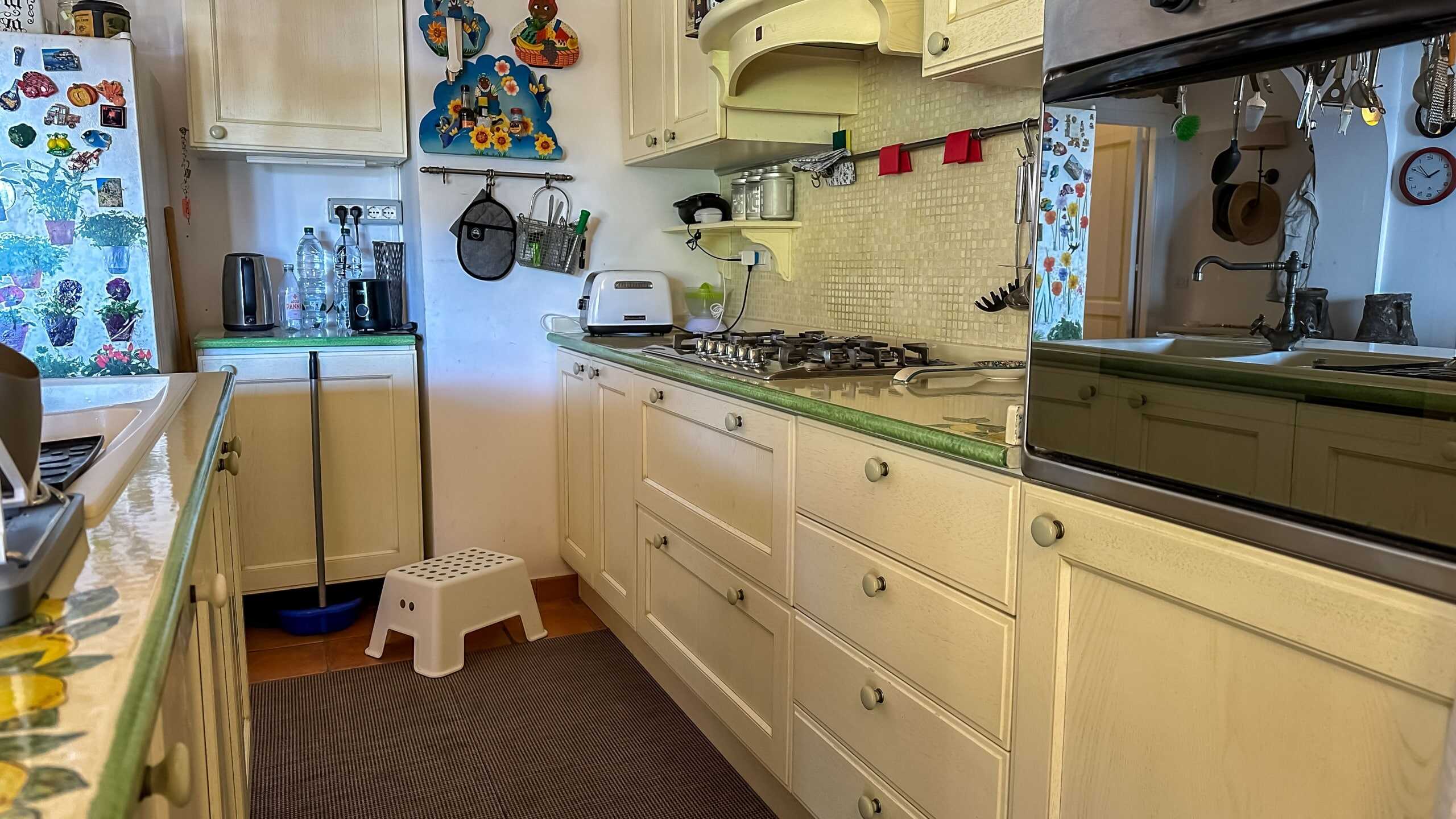 Lavagna cucina rustica vintage con mobili bianchi e dettagli colorati, ideale per case di campagna o bungalow, con elettrodomestici come forno e piano cottura, decorazioni floreali e spaziosa area cucina moderna per la vita quotidiana.