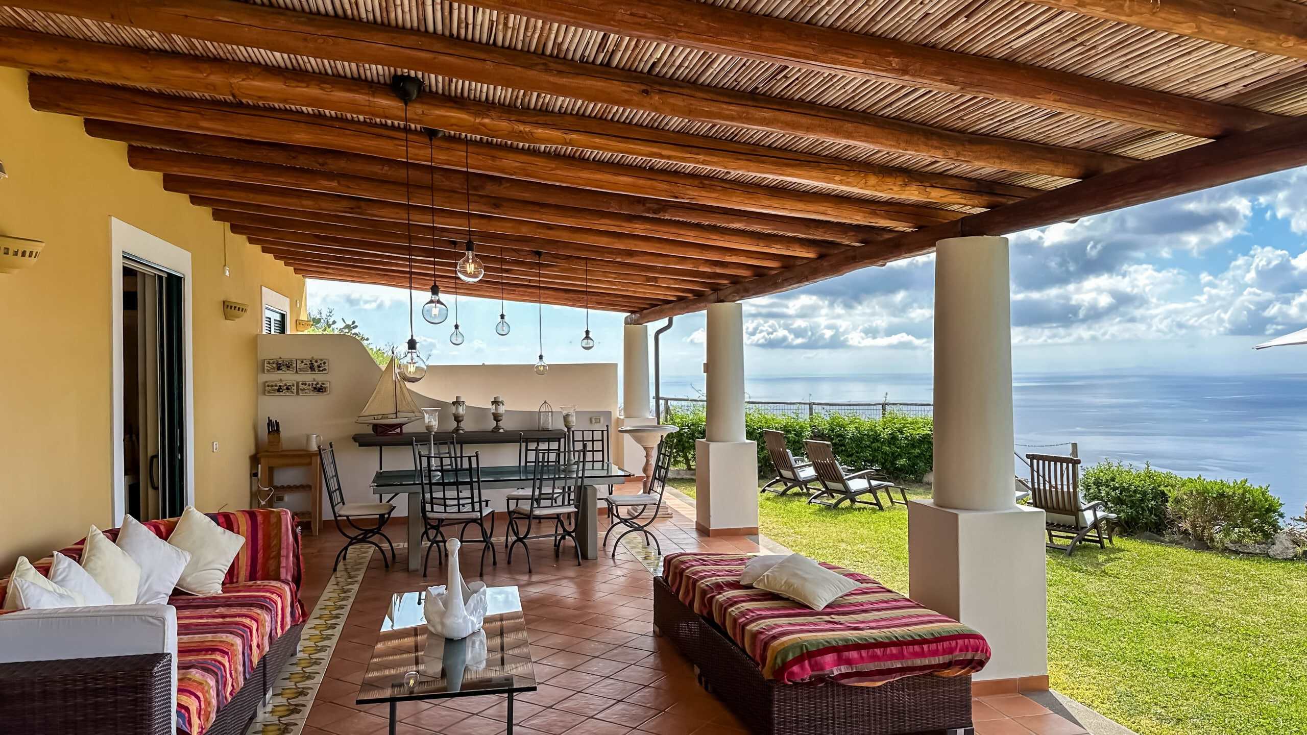 Relaxing outdoor living area with sea view, wicker furniture, wooden ceiling, and garden loungers, ideal for enjoying the Mediterranean climate and scenic vistas.