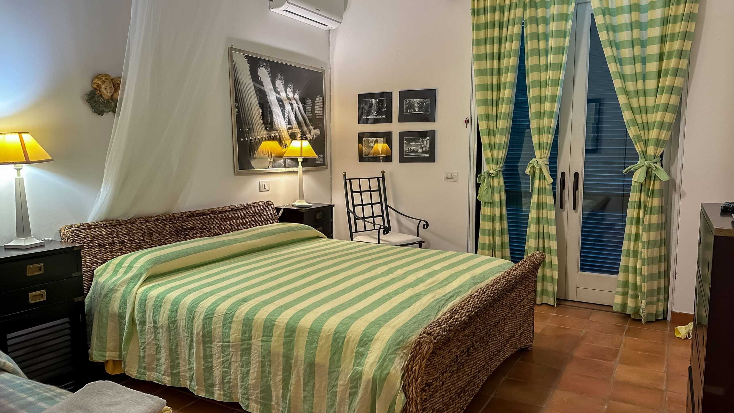L'immagine mostra una camera da letto con arredamento classico, con letto in stile rustico e tende a quadri verdi. La stanza è illuminata da lampade gialle, decorata con quadri e accessories che creano un ambiente accogliente e caldo. Perfetta per soggiorno in un contesto residenziale o turistico.