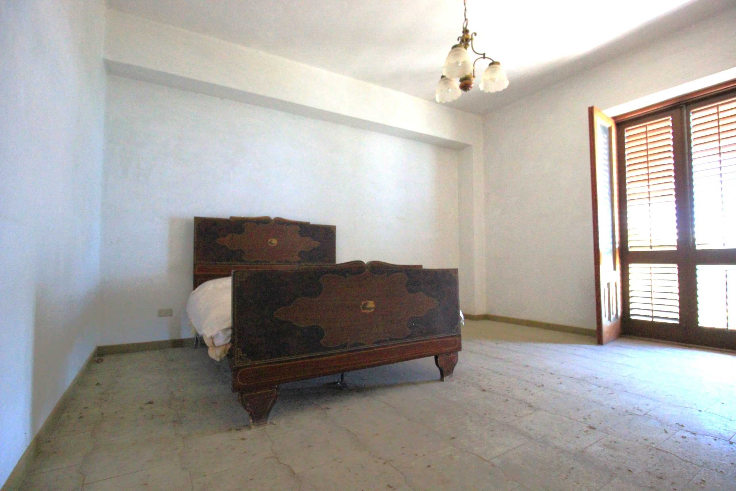 Vecchio letto in legno scuro in una stanza luminosa con pareti bianche e finestra a doppia anta con tapparelle di legno chiaro, illuminata dalla luce naturale.