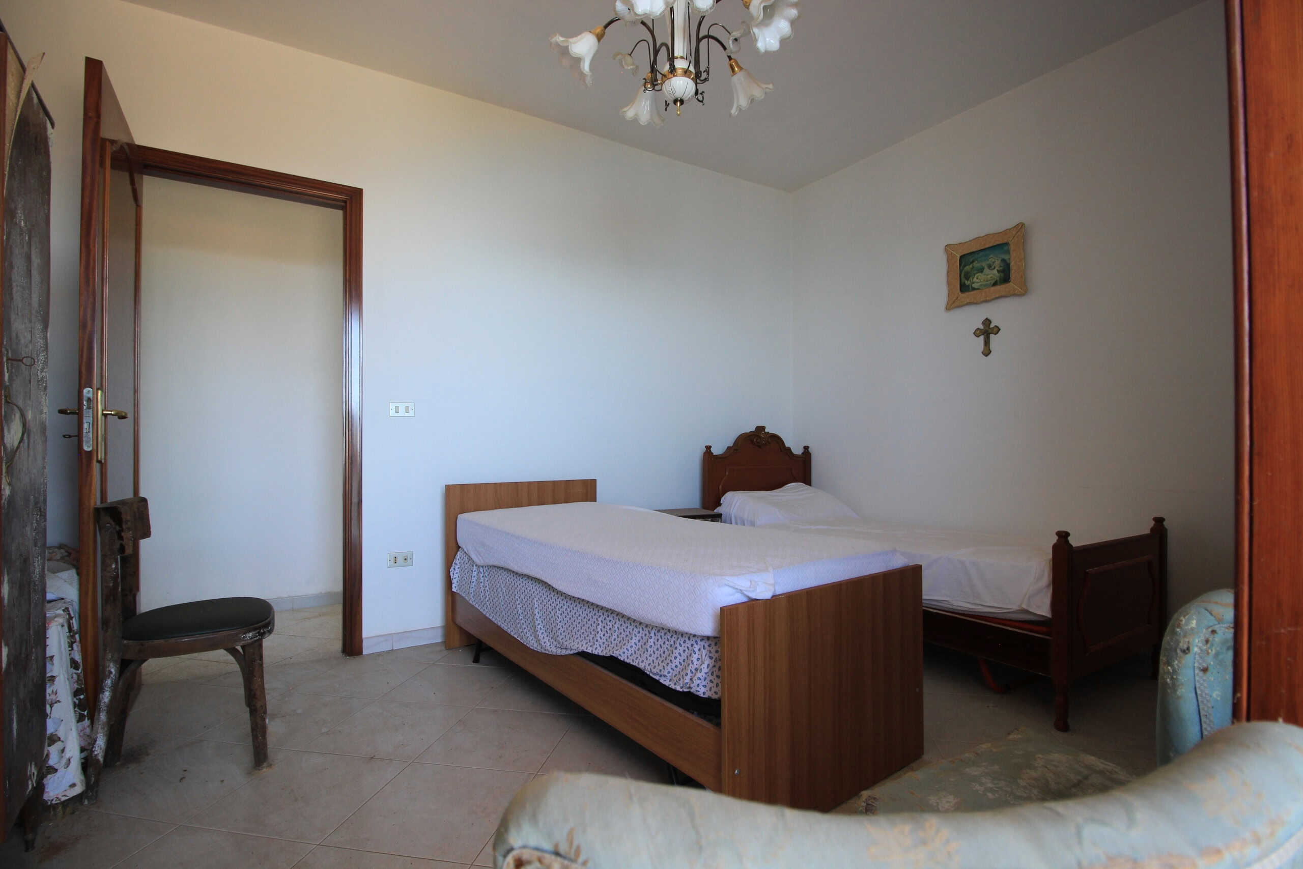 Letto singolo e doppio in stanza con arredi in legno, pareti bianche e decorazioni religiose, in ambiente immobiliare di NextCasa.