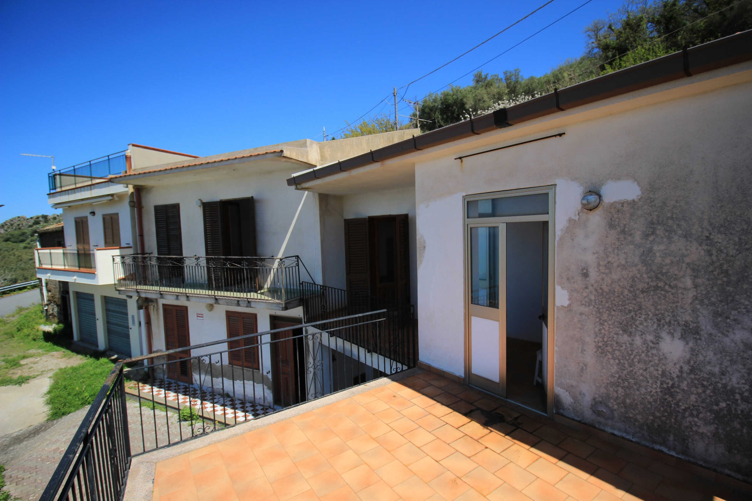 Balcone di un appartamento in stile mediterraneo, vista panoramica sulla natura e colline circostanti, zona abitativa con pavimentazione in piastrelle e finestre con persiane in legno.