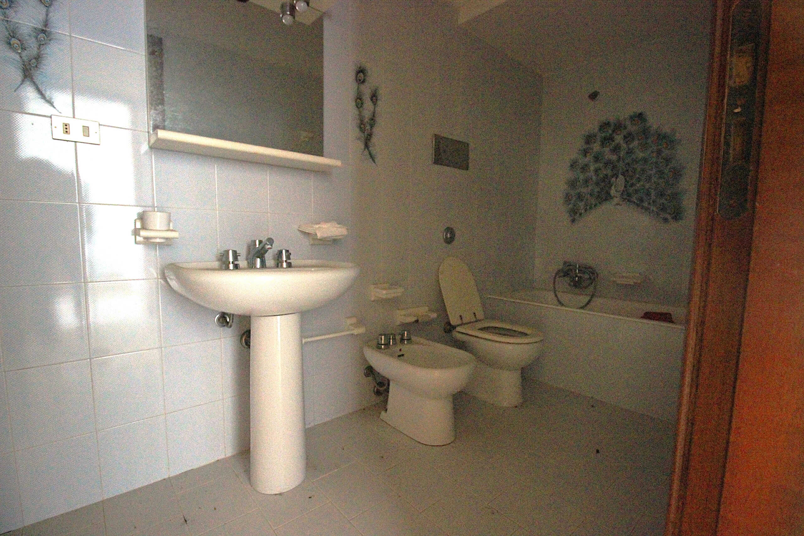 Lavabo e vater in bagno con piastrelle bianche, specchio e decorazioni, ambiente spazioso e luminoso adatto a soluzioni immobiliari di NextCasa.