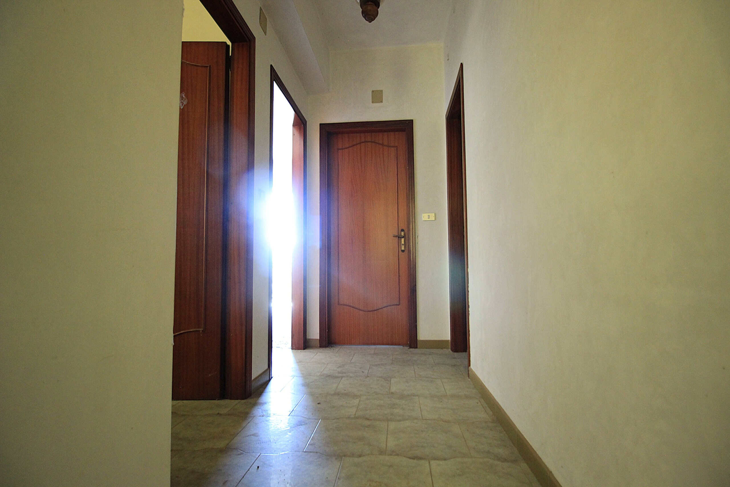 Porta d'ingresso in alluminio e legno con luce naturale che entra dall'apertura aperta, in un appartamento o casa. La parete è dipinta di colore beige chiaro, con pavimento in gres porcellanato. La scena rappresenta un ingresso o corridoio interno, con porte in legno e una porta scorrevole in vetro e legno sul lato sinistro. L'ambiente è semplice, funzionale, ideale per abitazioni in Toscana o in zone residenziali.