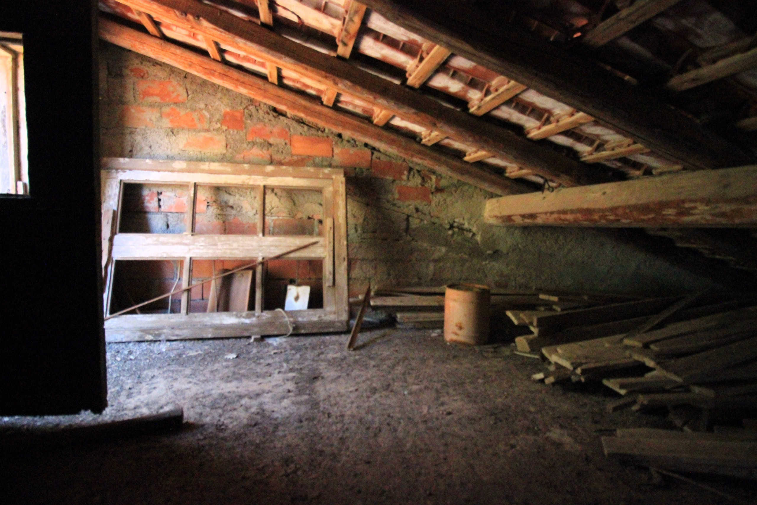 Vecchia soffitta in fase di ristrutturazione, con pareti in mattoni e pavimento di terra, elementi in legno e tegole apparenti, pronta per essere trasformata in uno spazio abitabile o deposito.