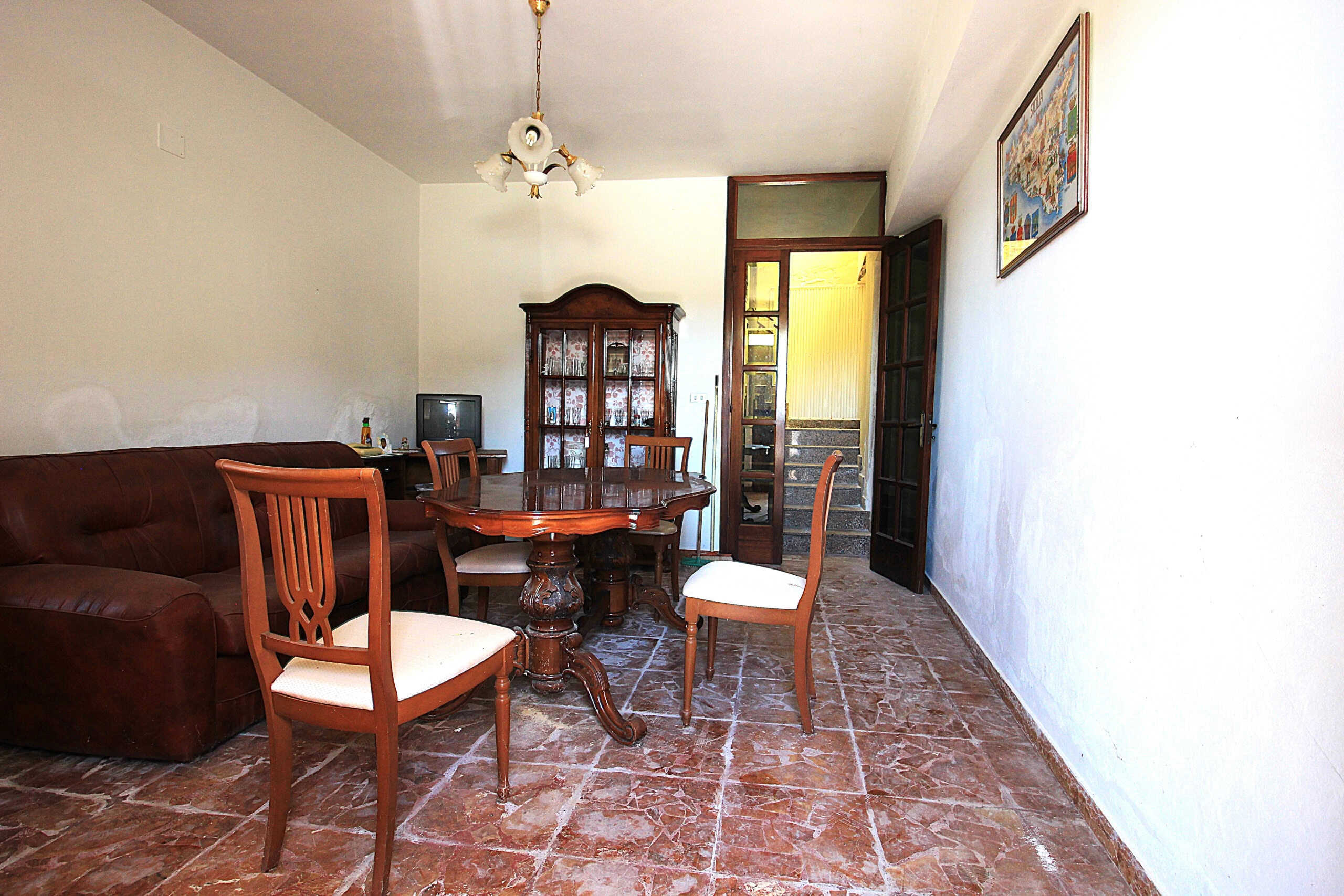 Elegante soggiorno con pareti bianche, pavimento in pietra, tavolo in legno e sedie imbottite, arredi classici, credenza in legno e TV, ideale per case in vendita o affitto a NextCasa.