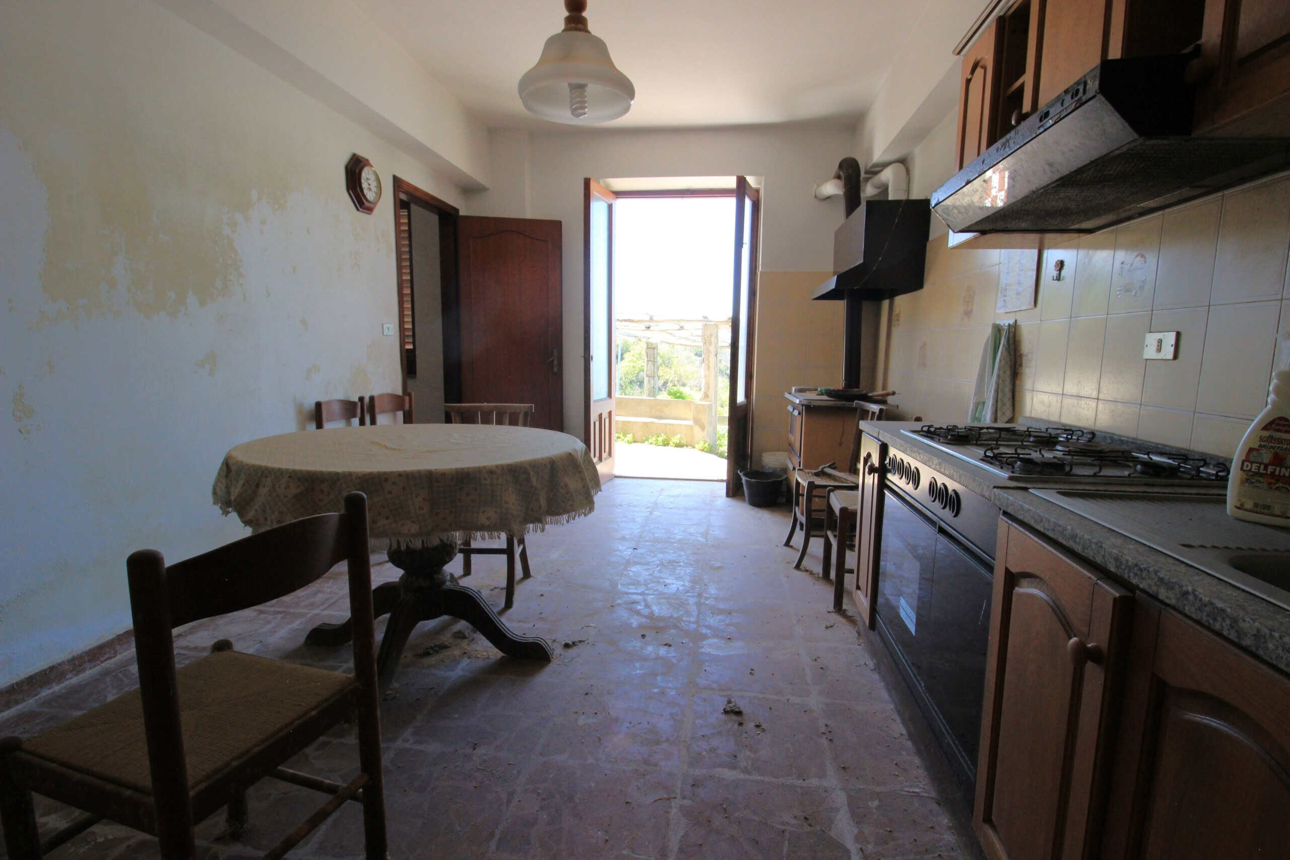 Vecchia cucina rustica con tavolo rotondo, sedie e cucina tradizionale in stile country, ambiente luminoso con porta verso vista panoramica, ideale per affitto case vacanza NextCasa.