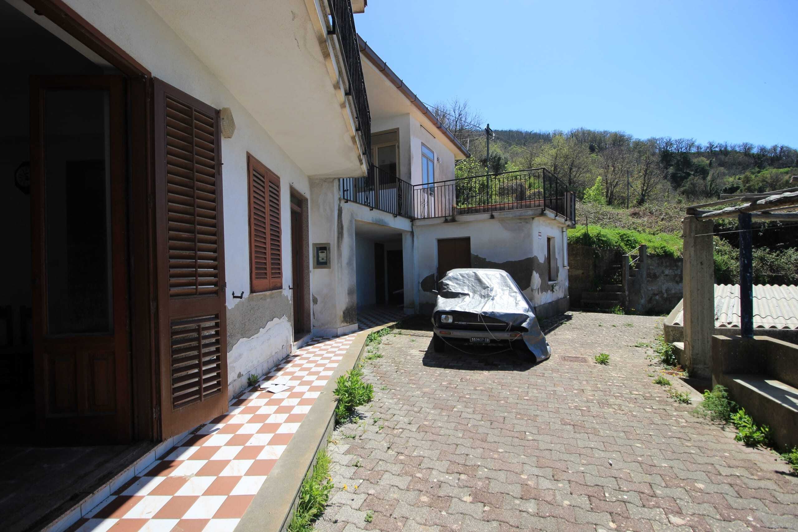 Automobile parcheggiata davanti a casa in zona rurale, edificio residenziale con finestre con persiane di legno, spazio esterno con piante e vista su colline e vegetazione, ideale per promuovere meravigliose case in campagna.