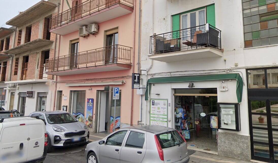 Fronte di un edificio residenziale con negozio al piano terra, parcheggio stradale con auto, dettagli di balconi, aria condizionata, e insegne commerciali in zona urbana.