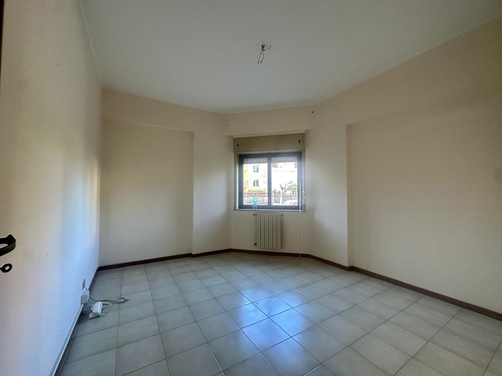 Ampio ambiente vuoto con una grande finestra, pavimento in piastrelle chiare, pareti neutre e radiatore, perfetto per l'acquisto di immobili con servizi immobiliari NextCasa.
