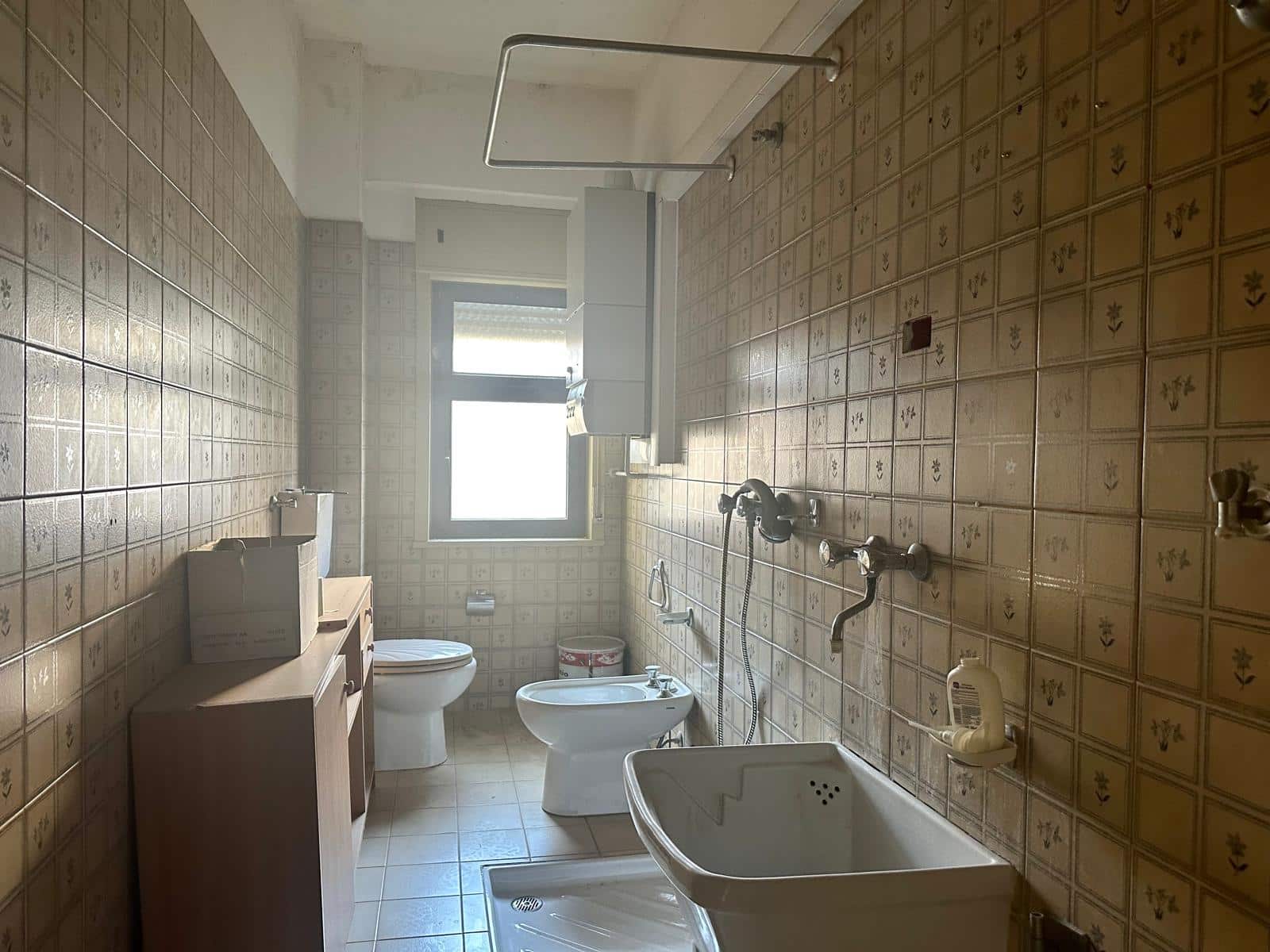 Bagno con sanitari, bidet e vasca in stile vintage, pareti rivestite con piastrelle beige con fiori, finestra naturale, arredi semplici e funzionali, ideale per ristrutturazione o rinnovamento domestico.